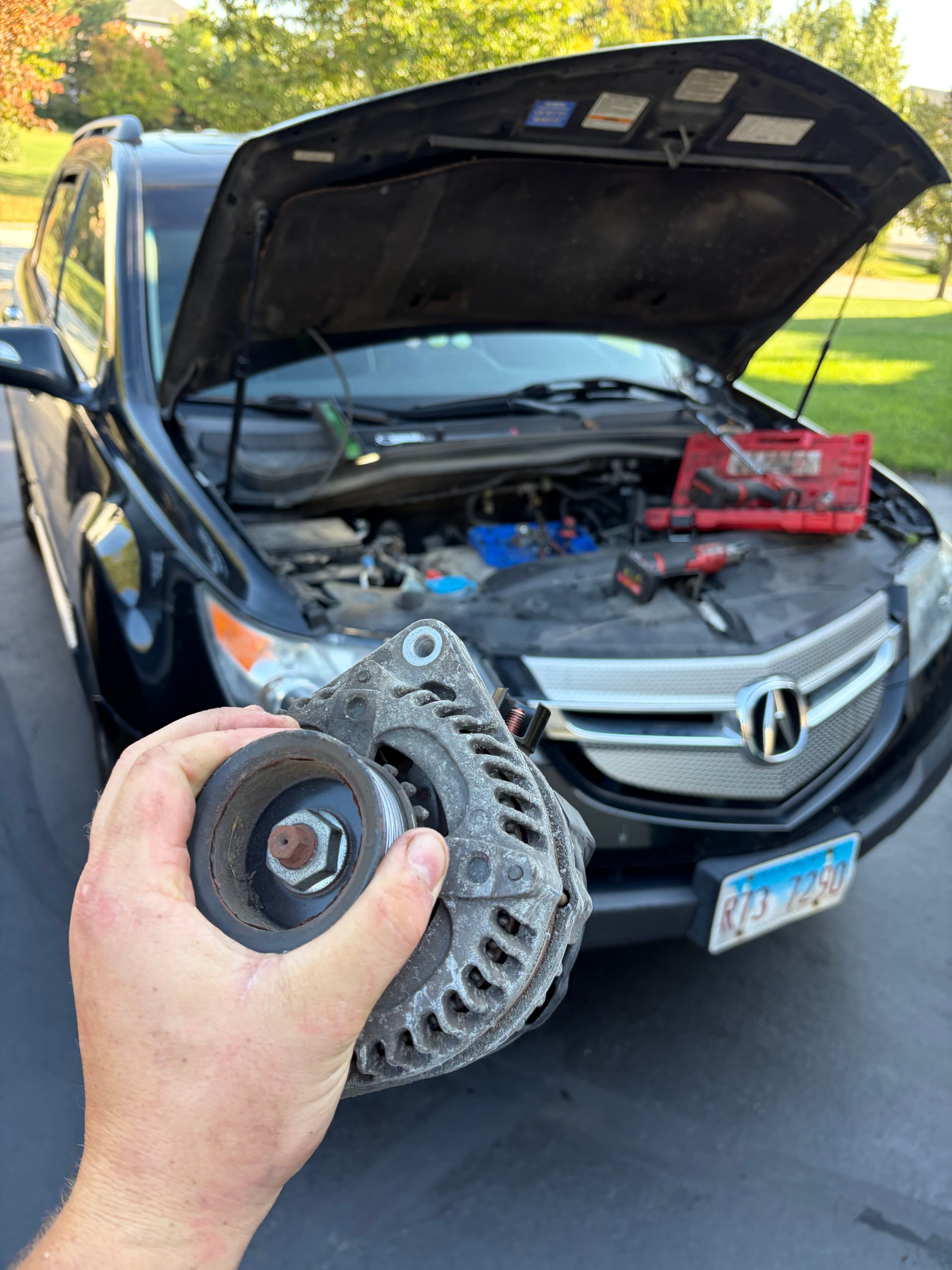 Hand holding a car alternator in front of an open hood with tools visible.