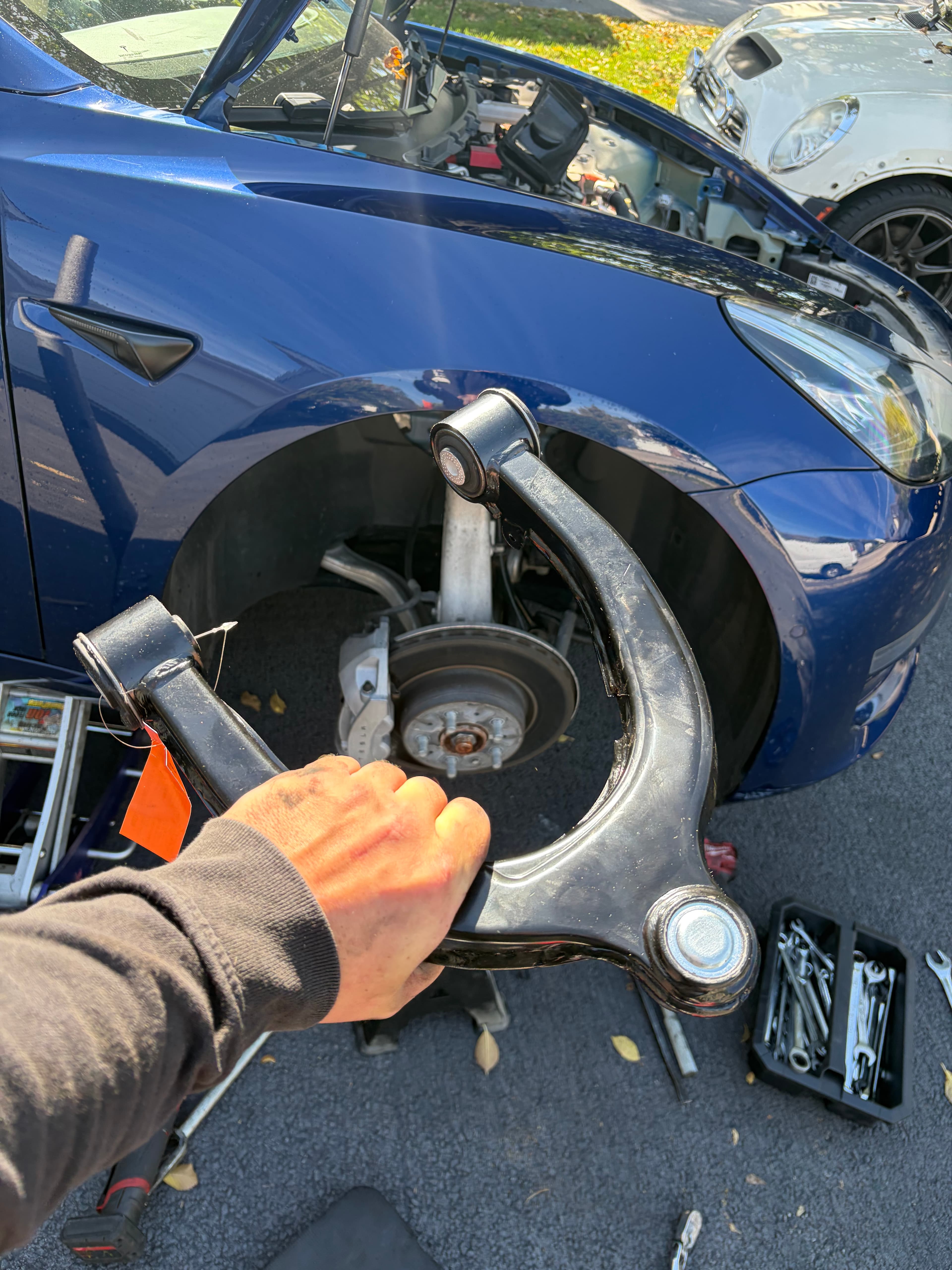 A hand holds a black upper control arm over a blue car's wheel assembly.