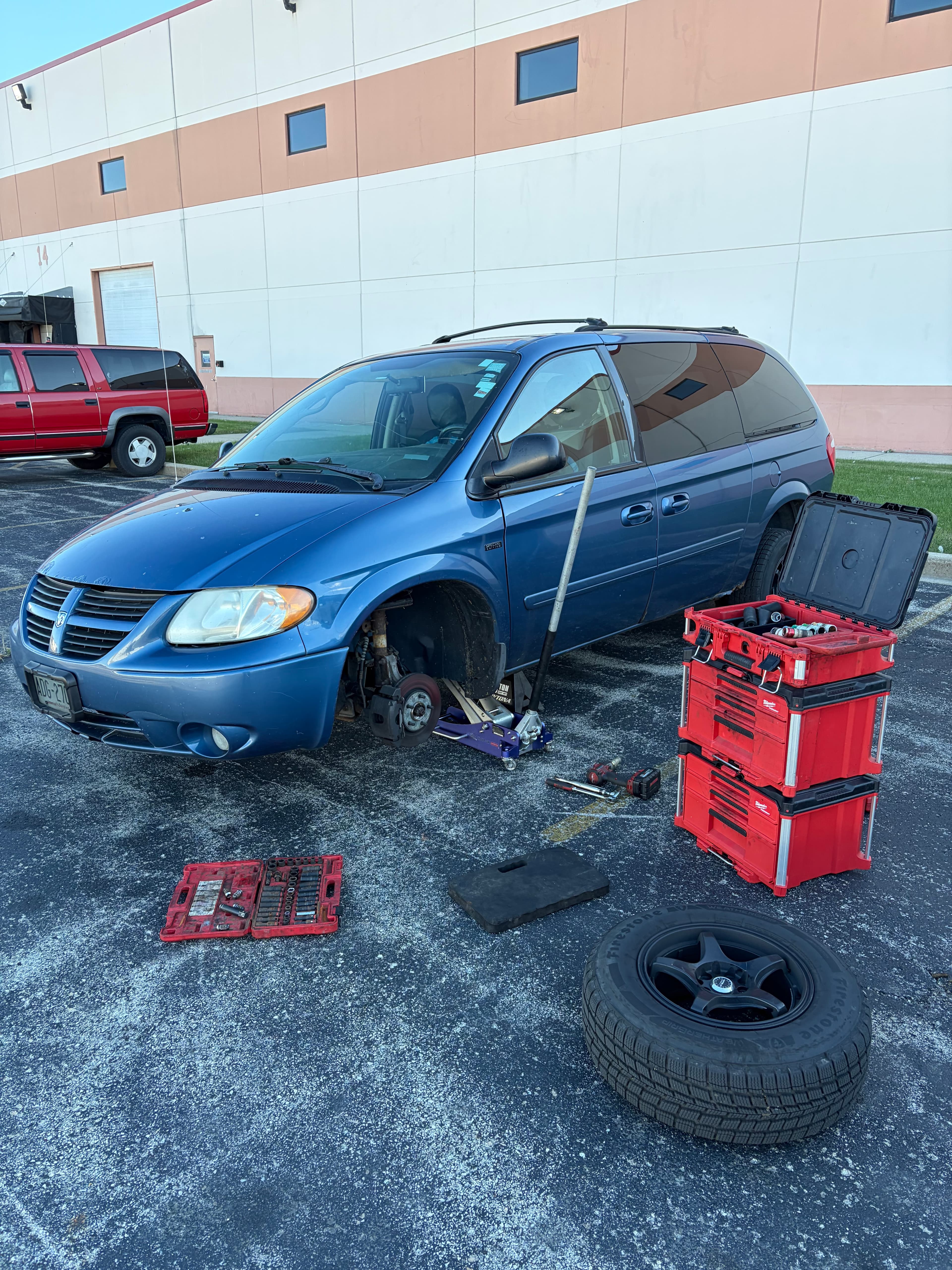 Blue minivan jacked up in a parking lot with wheel removed and red toolboxes.
