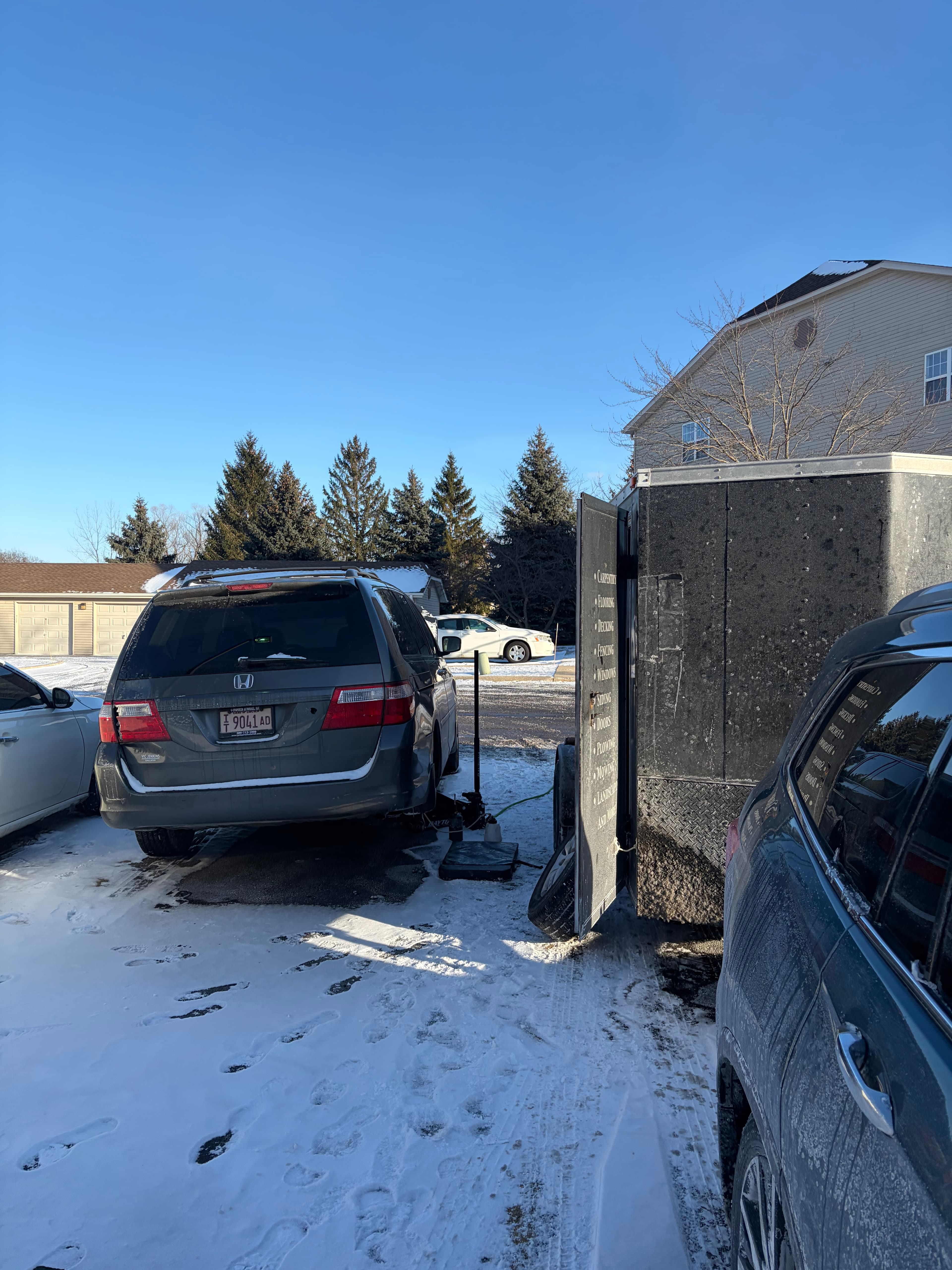 Grey Honda minivan parked in a snowy driveway next to a large black utility trailer.