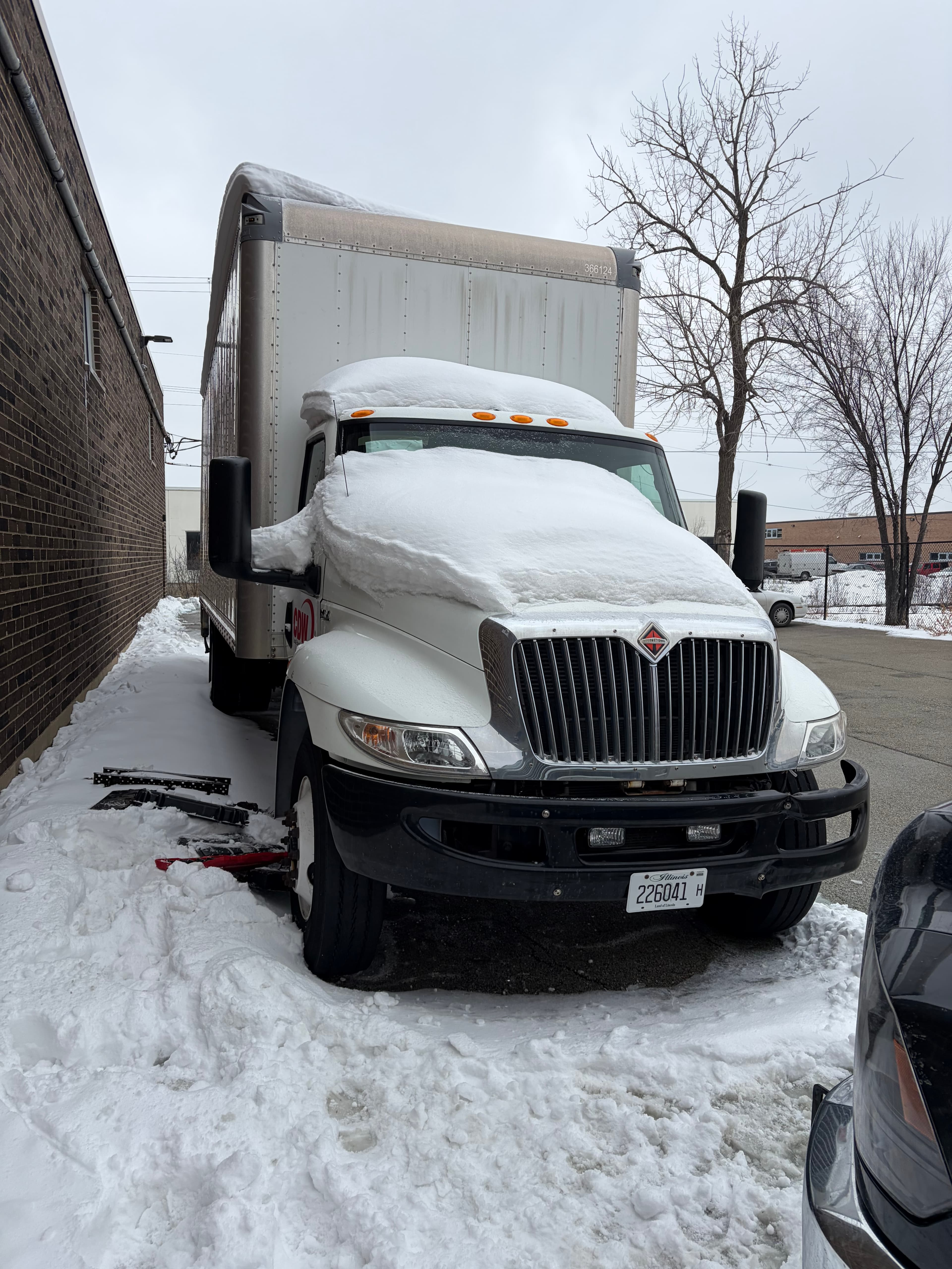 White box truck buried under a thick layer of snow next to a brick building.