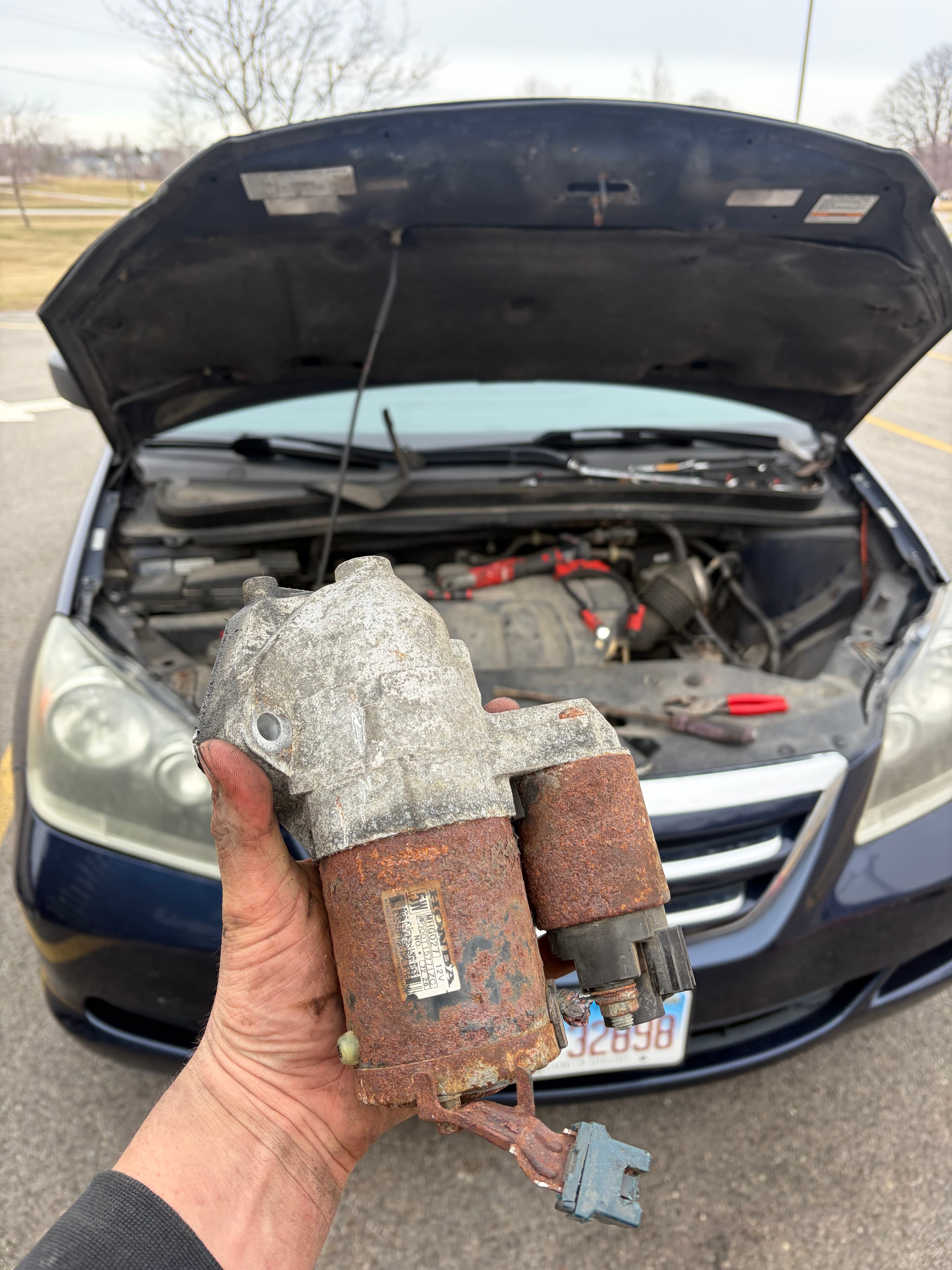Hand holding a rusty Honda starter motor in front of a car's open hood.