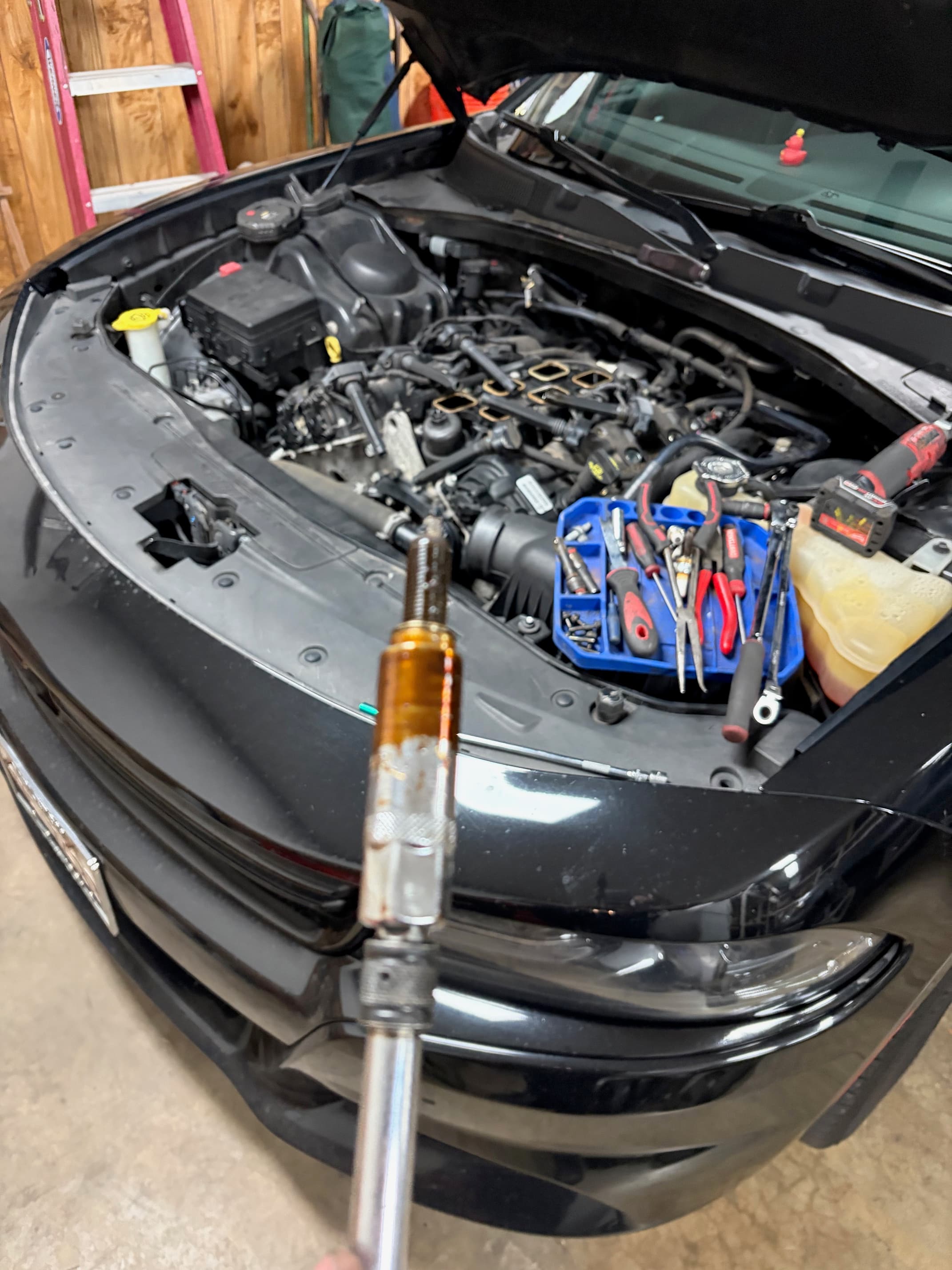 Oil-soaked spark plug held over an open car engine bay with various repair tools.