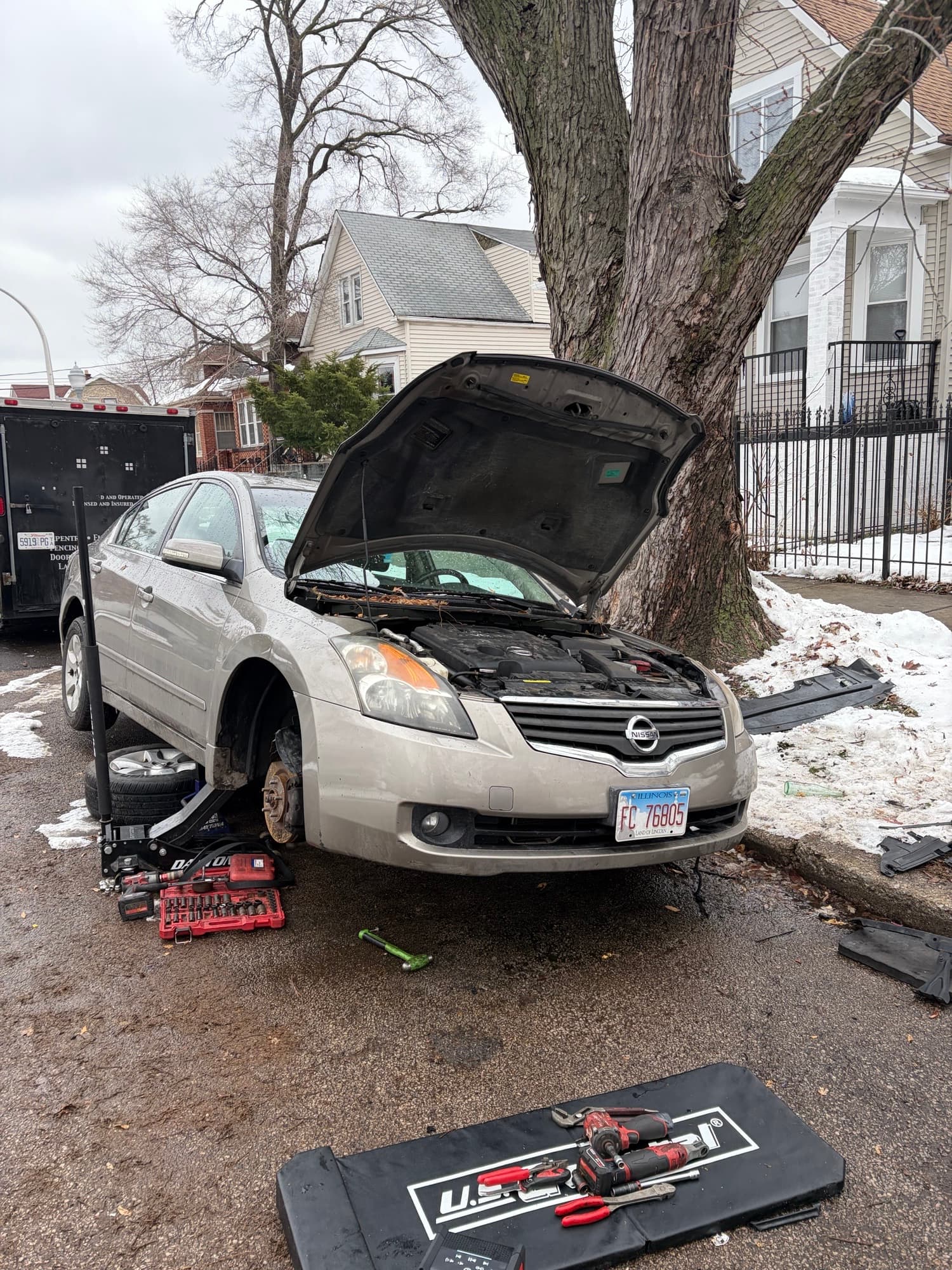 Silver Nissan car with open hood jacked up on a driveway with mechanic tools.